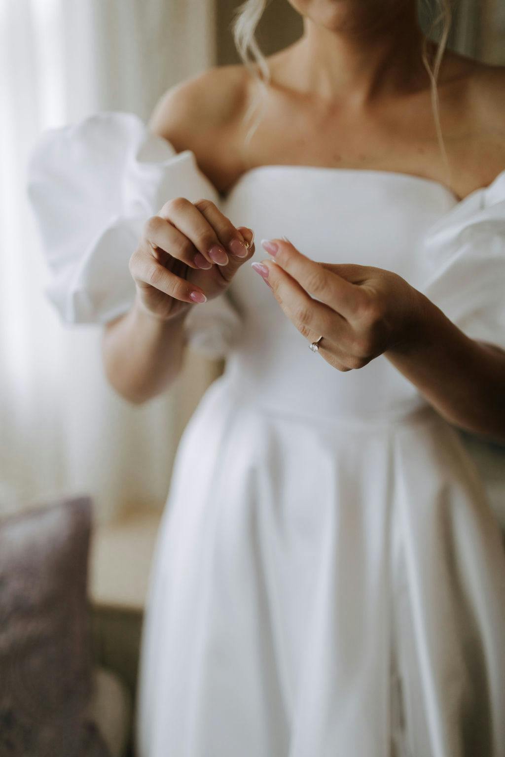 A bride in a white off-the-shoulder wedding dress with puffy sleeves is seen from the neck down. She is holding a ring in her hands, and there's a glimpse of a wedding ring on her finger. The background shows a softly lit room with a curtain and a piece of furniture.