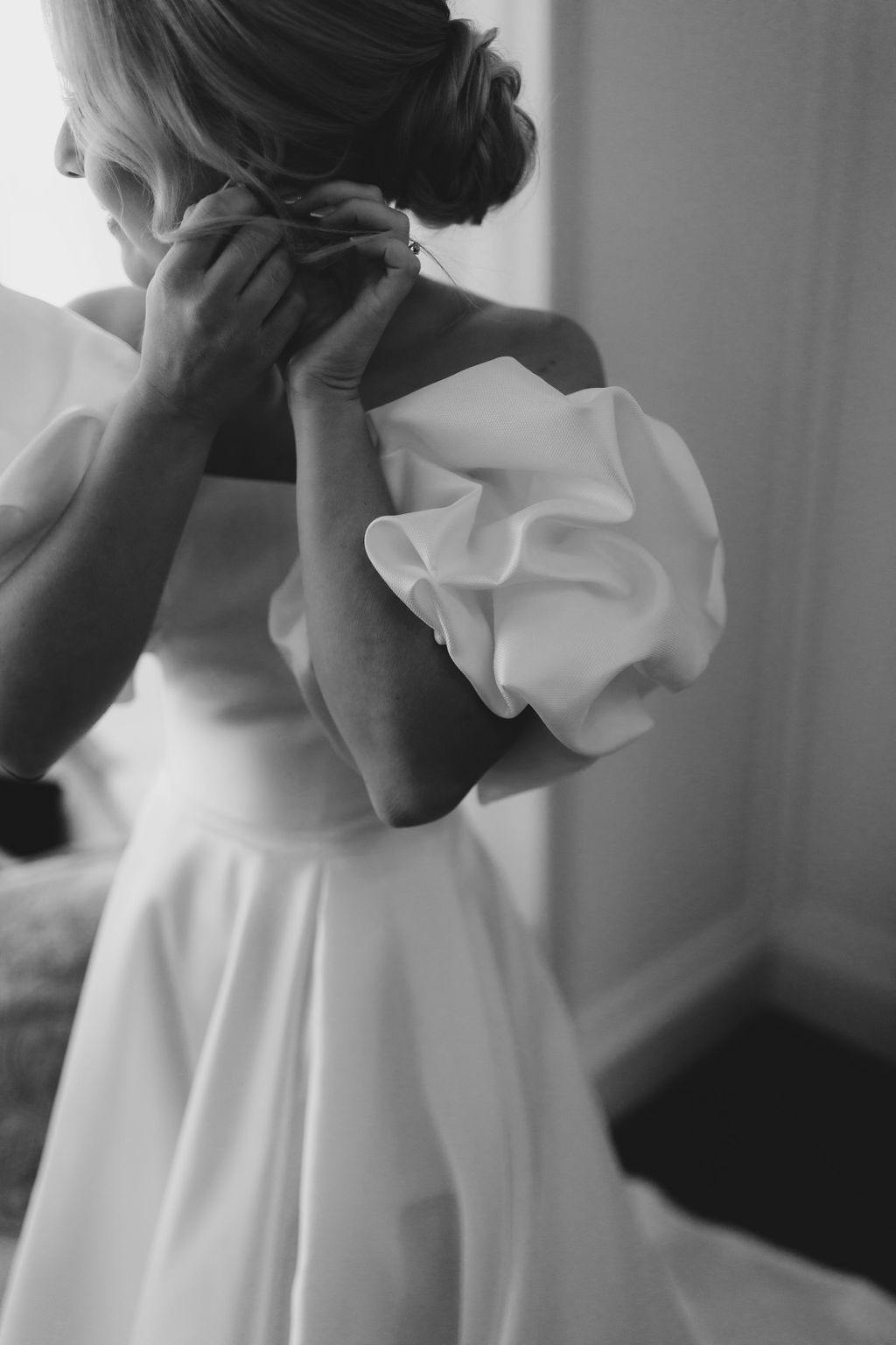 A black and white image of a person in a wedding dress. The dress features elegant, puffy sleeves and a fitted bodice. The person is adjusting an earring and looking away from the camera, with their hair styled in an updo. The background is softly lit.