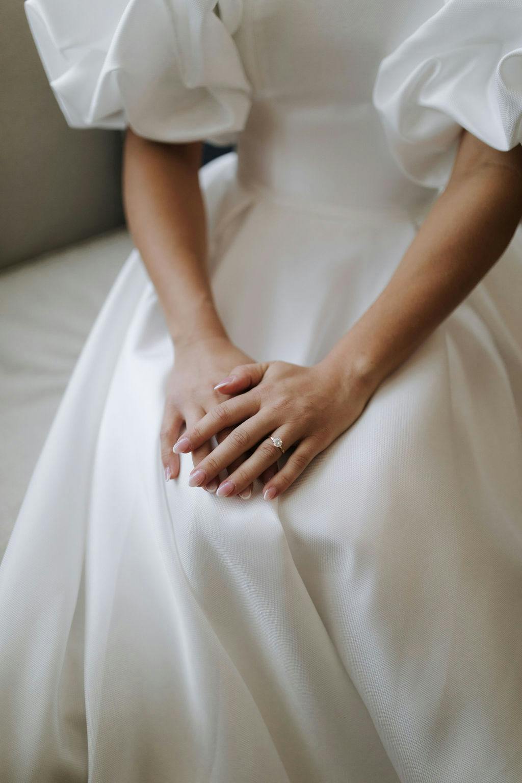 A bride wearing a white dress with puffed sleeves rests her hands on her lap, showcasing a simple yet elegant engagement ring on her finger.