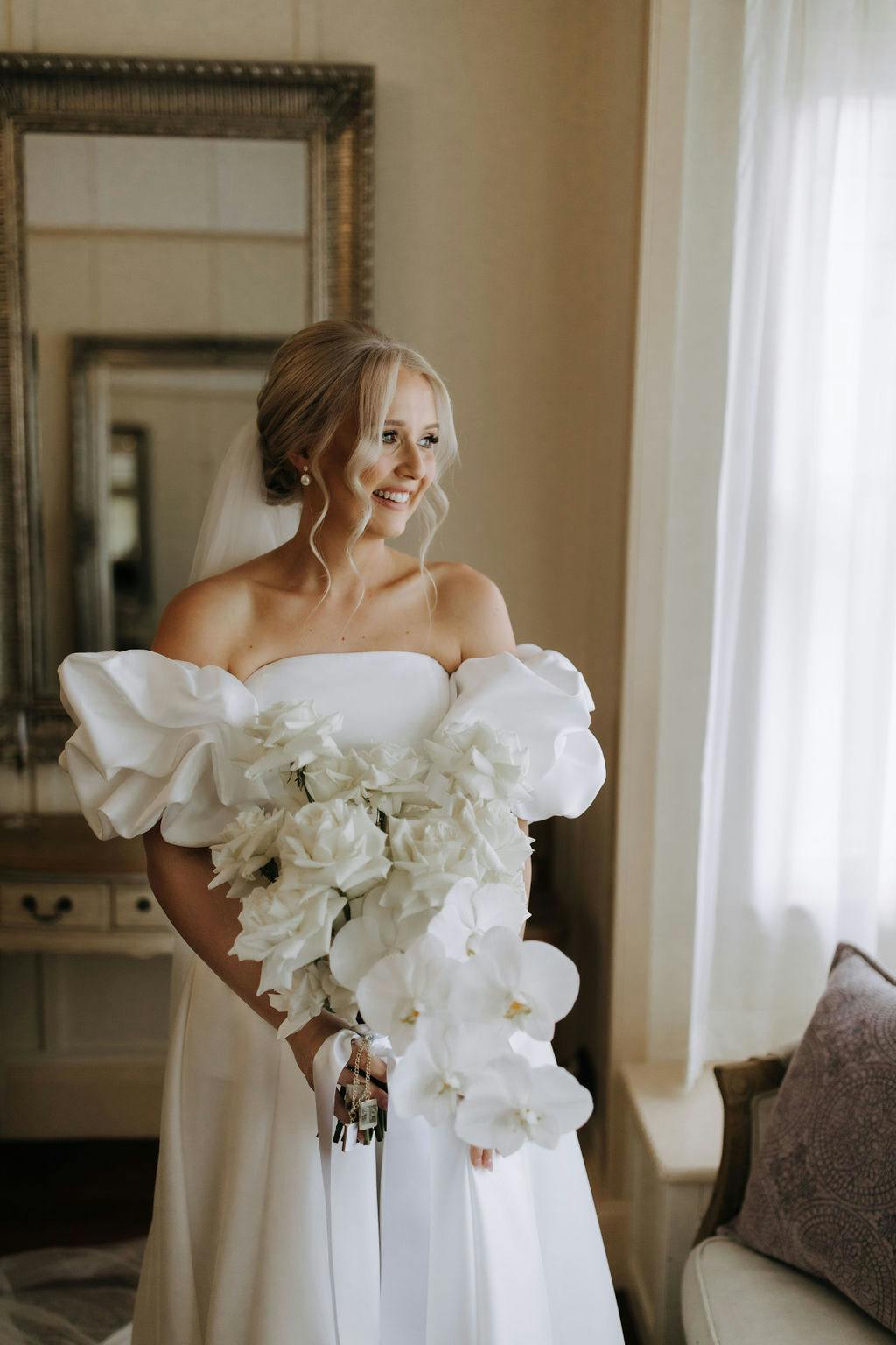 A bride with blonde hair stands indoors by a window with sheer curtains. She wears an off-the-shoulder white wedding dress with puffy sleeves and holds a large bouquet of white flowers, smiling and looking toward her left. A mirror and a small desk are in the background.