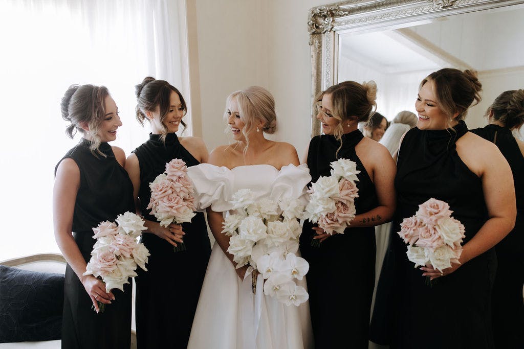 A bride in a white dress stands smiling with four bridesmaids wearing black dresses. Each woman holds a bouquet of white and pink flowers. They are standing in a room with a large mirror and a window with sheer white curtains.