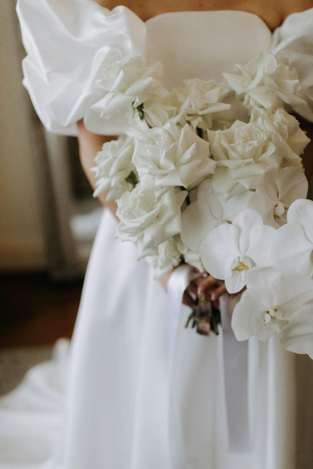 A bride in a white gown holds a bouquet of white roses and orchids, with puffed sleeves of her dress visible. The background is softly blurred, focusing the attention on the flowers and the fabric details of the dress.