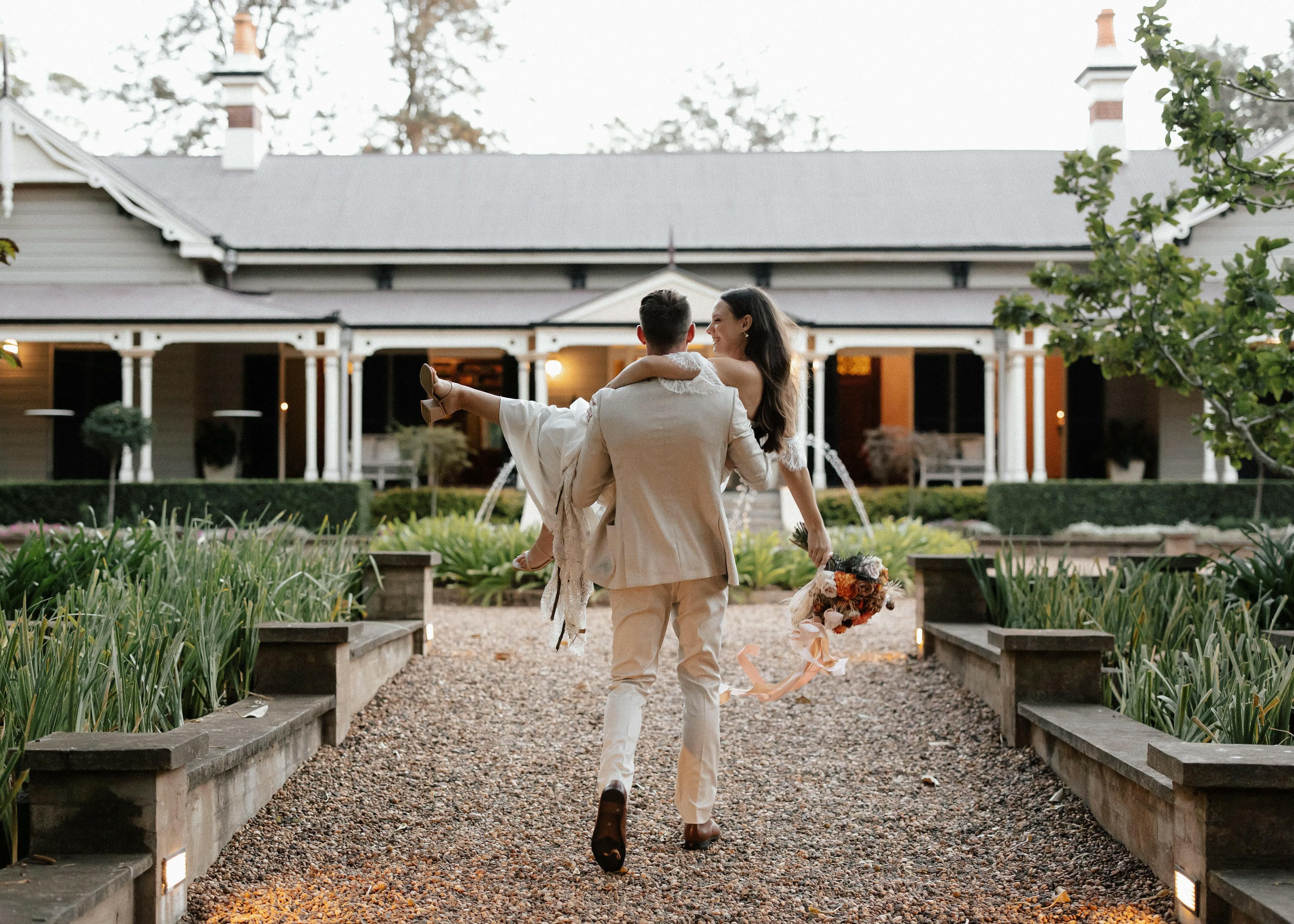 A groom carrying his bride in front of an elegant, rustic building with a garden. He is walking on a gravel path, both dressed in wedding attire; the bride holding a large bouquet of flowers. The background showcases lush greenery and a charming wooden facade.