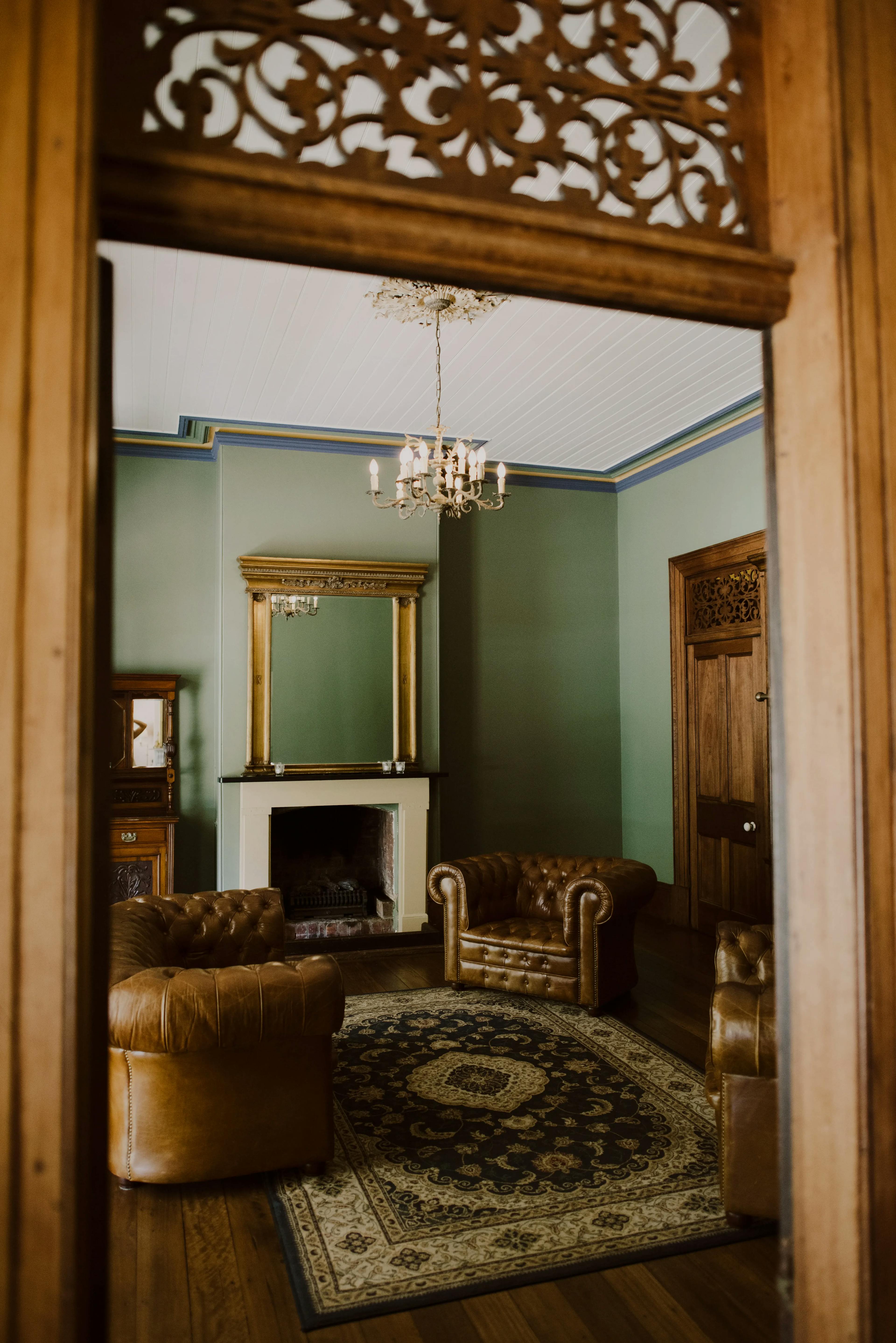 Cozy room with green walls viewed through a carved wooden frame. It features a chandelier, ornate fireplace with a large mirror above it, and three brown leather chairs around a patterned rug on a wooden floor.