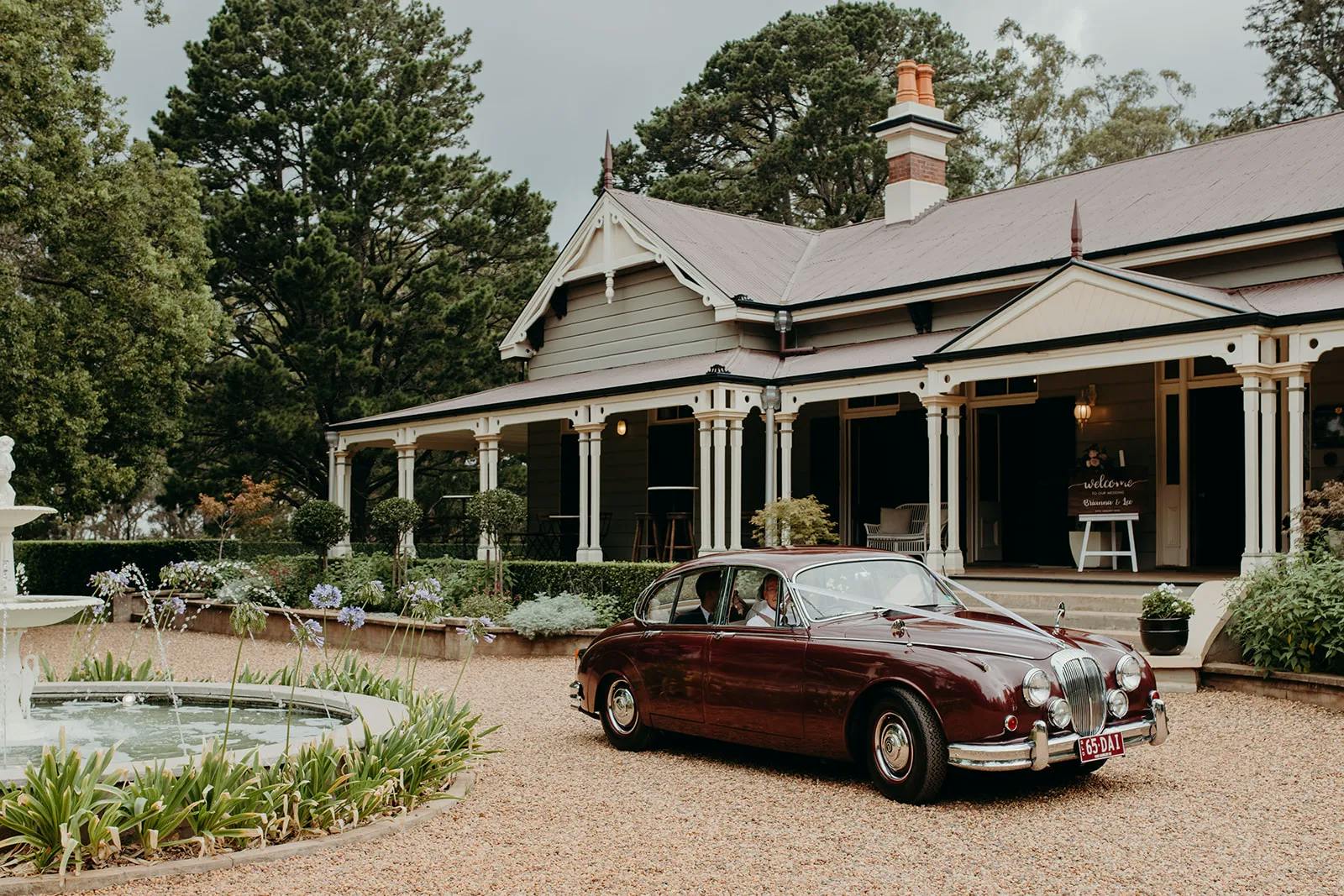 A classic, maroon vintage car is parked on a gravel driveway in front of an elegant, large house. The house features a wraparound porch, ornate trim, and a manicured garden with a fountain in the foreground. Tall trees surround the property under a cloudy sky.