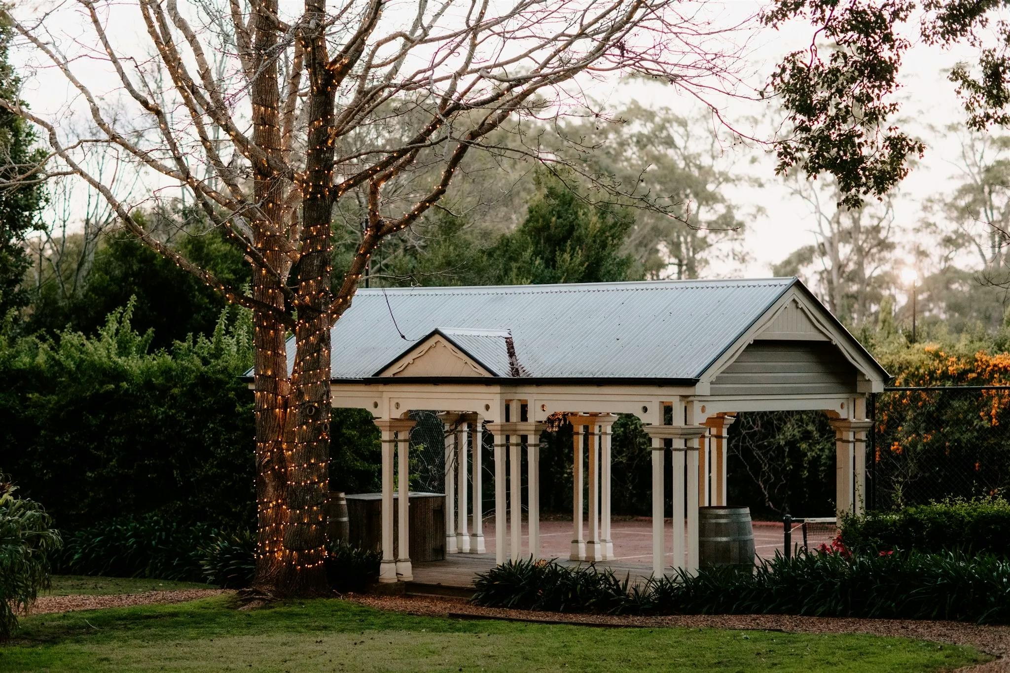 A quaint outdoor pavilion stands with a sloped, grey roof, flanked by slender white columns. The adjacent large tree is wrapped with string lights, creating a cozy atmosphere. The scene is surrounded by lush greenery on a peaceful day.