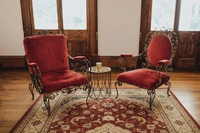 The image shows two ornate, vintage-style red velvet armchairs with intricate metal frames facing each other. Between them is a small round metal table with a glass of water on top. The scene is set on a patterned area rug, with wooden doors and windows in the background.