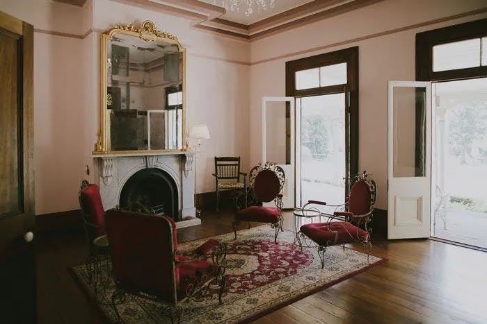A stylish room featuring a large mirror above a white fireplace with a decorative mantle. There are four ornate chairs with red upholstery arranged on a patterned rug. The room has large windows and glass doors that open to a garden.
