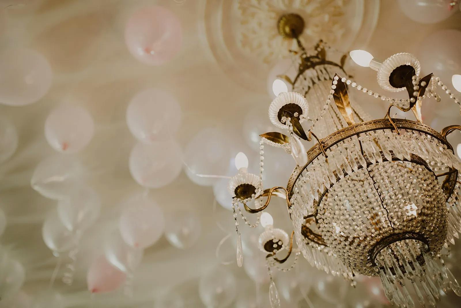 An ornate crystal chandelier with lit bulbs hangs from a ceiling decorated with white and pink balloons. The chandelier features intricate details and gold accents, adding an elegant touch to the festive ambiance.