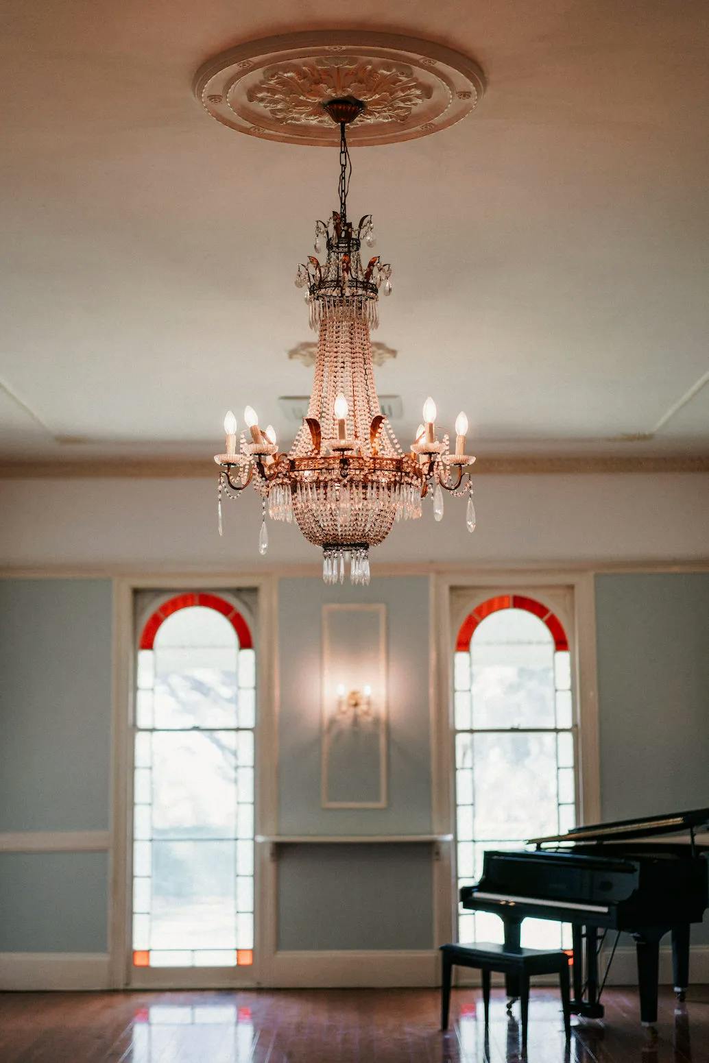 An elegant room with a grand crystal chandelier hanging from the ceiling. The room features large windows with red and white accents, illuminating the sparse space. In the corner, there is a black grand piano. The floor is polished wood.