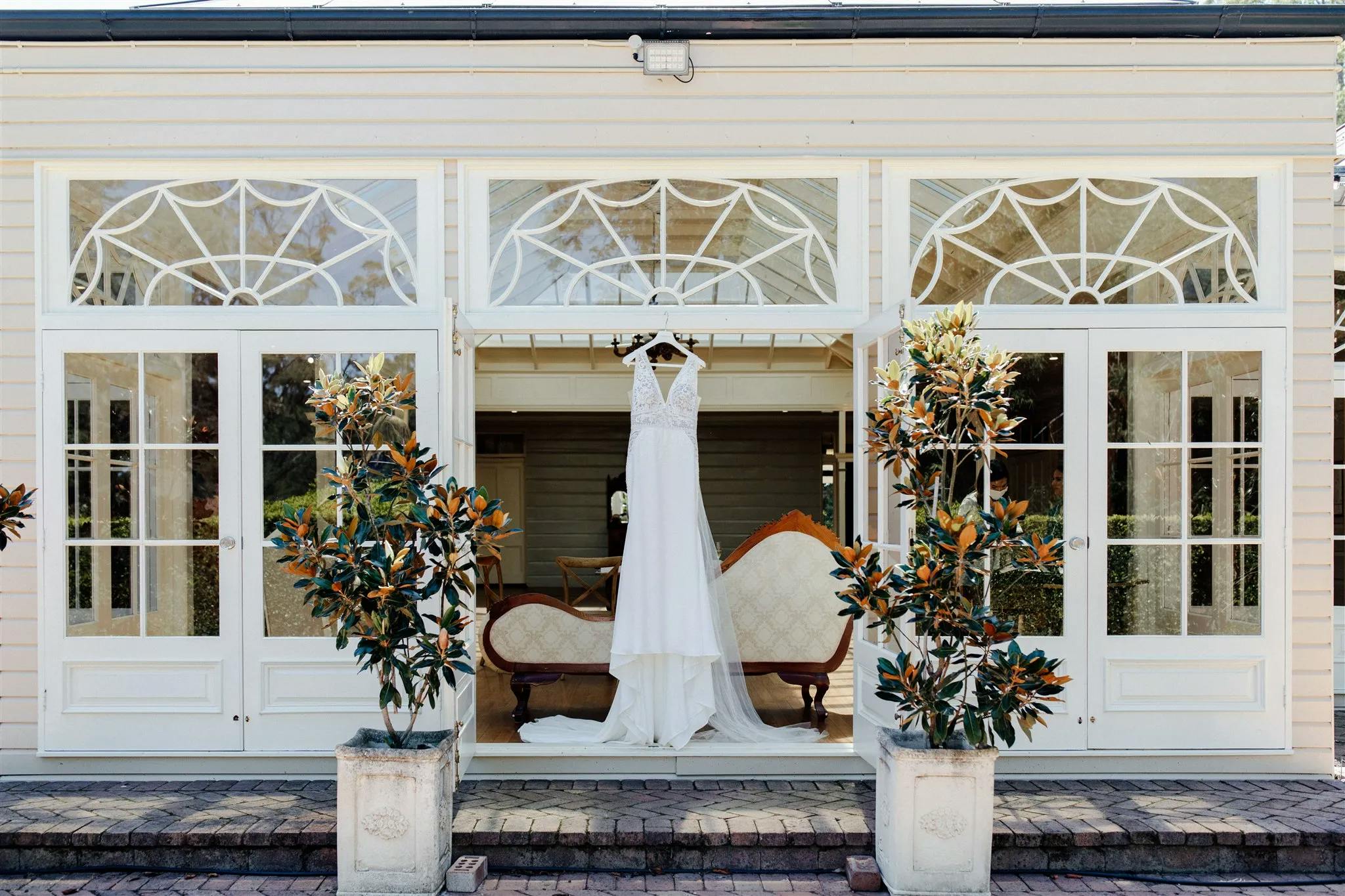 A white wedding dress hangs in front of a large elegant window. The window features multiple glass panes with decorative patterns. Two potted plants frame the entrance, and there is vintage furniture, including a sofa, visible inside the building.