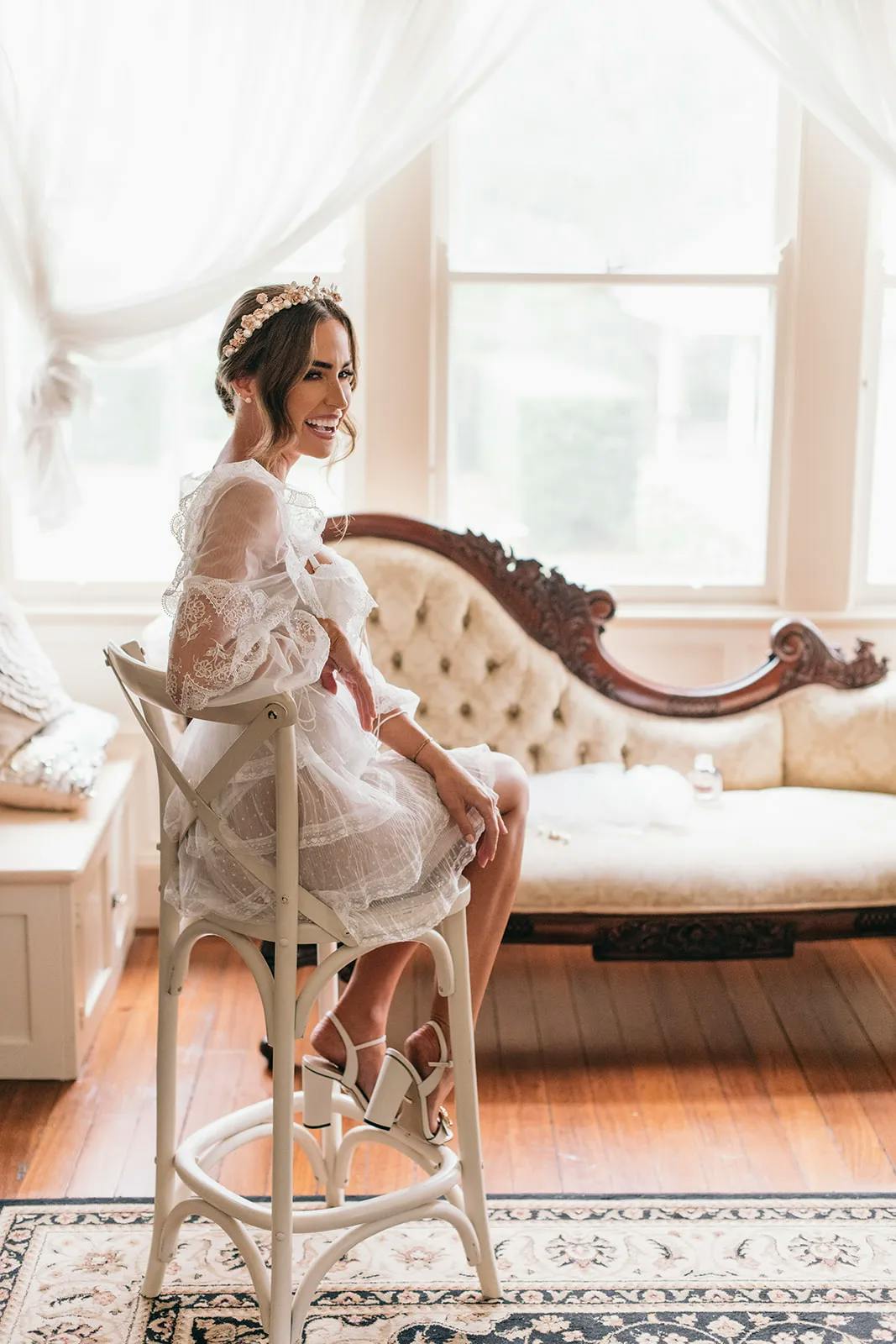 A woman in a white, lacy dress and a delicate floral headband sits on a wooden chair near a vintage sofa in a sunlit room. She smiles brightly, and the room has wooden floors and large windows draped with sheer curtains.