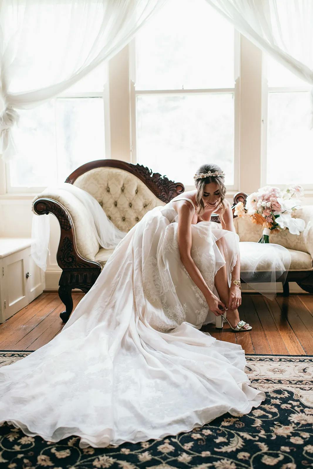 A bride in a white wedding dress sits on an elegant vintage sofa in a sunlit room with large windows. She adjusts her shoe, with a bouquet of flowers beside her on the sofa. The dress flows onto the wooden floor, and sheer curtains adorn the windows.