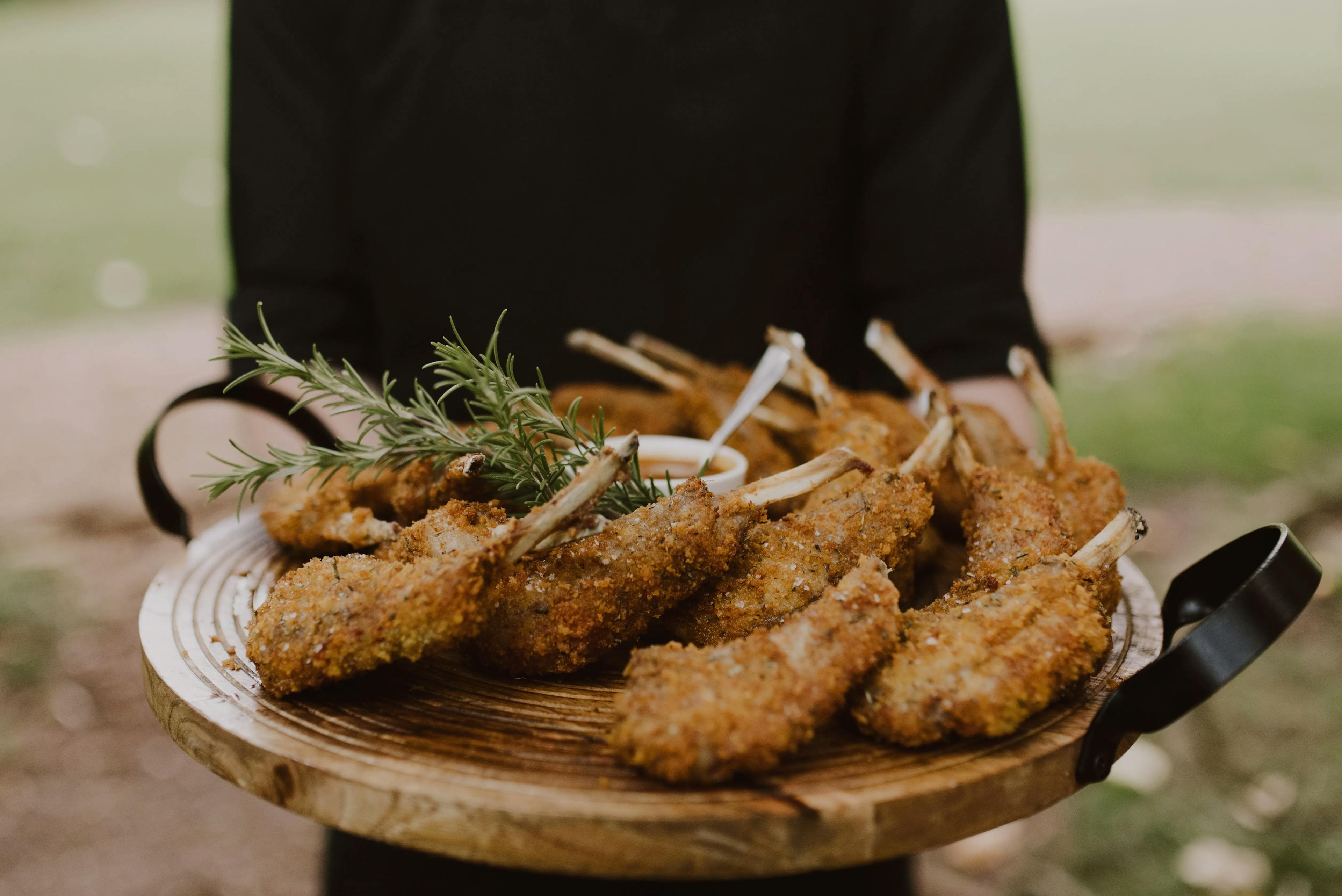 A person holding a round wooden serving board with a variety of breaded and fried lamb chops, garnished with sprigs of rosemary and accompanied by a small bowl of dipping sauce, set against a blurred outdoor background.