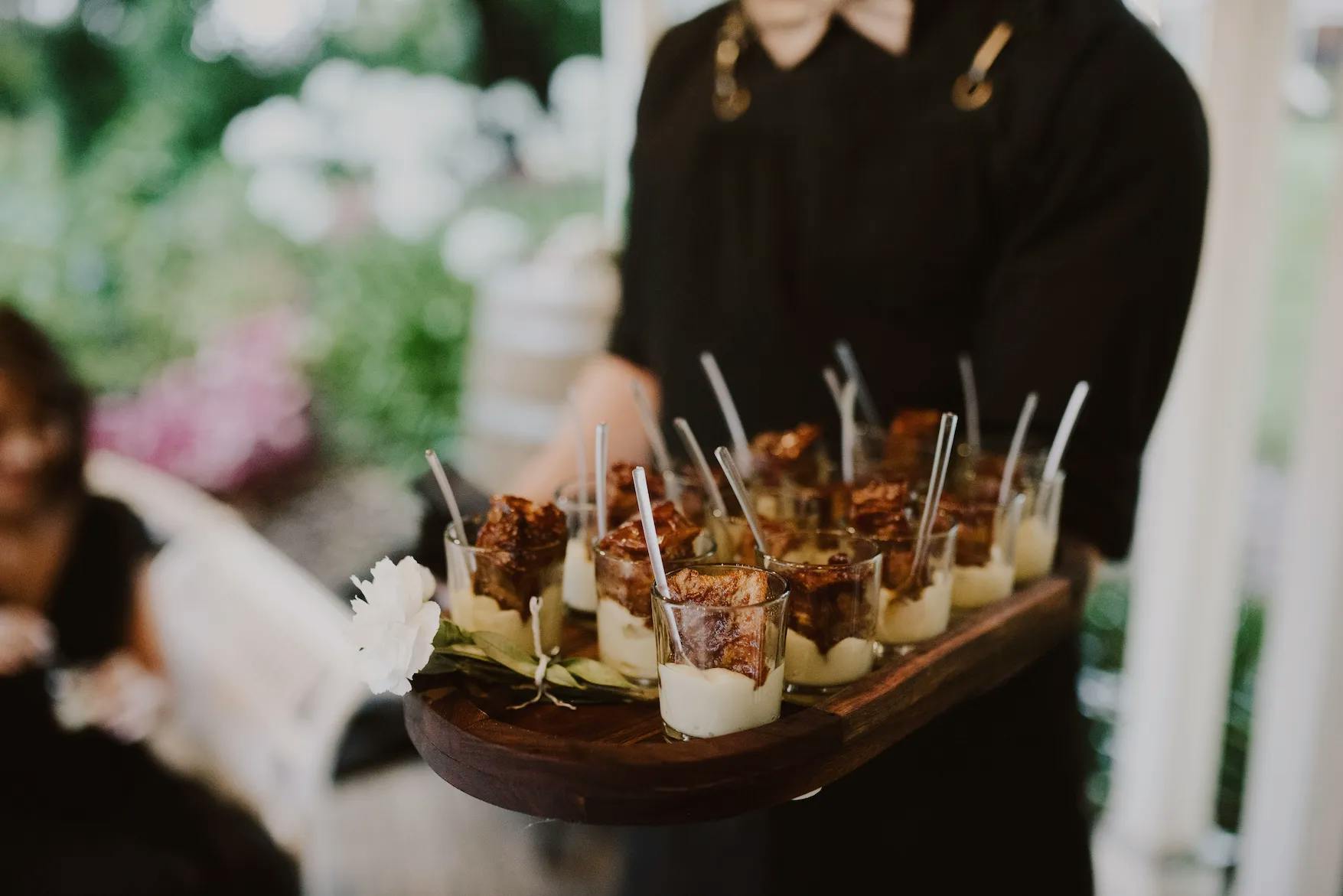 A person in a black outfit holds a wooden tray with several small dessert cups. Each cup contains a creamy dessert topped with a caramelized crust and a small spoon. In the background, there's an outdoor setting with greenery and blurred figures.
