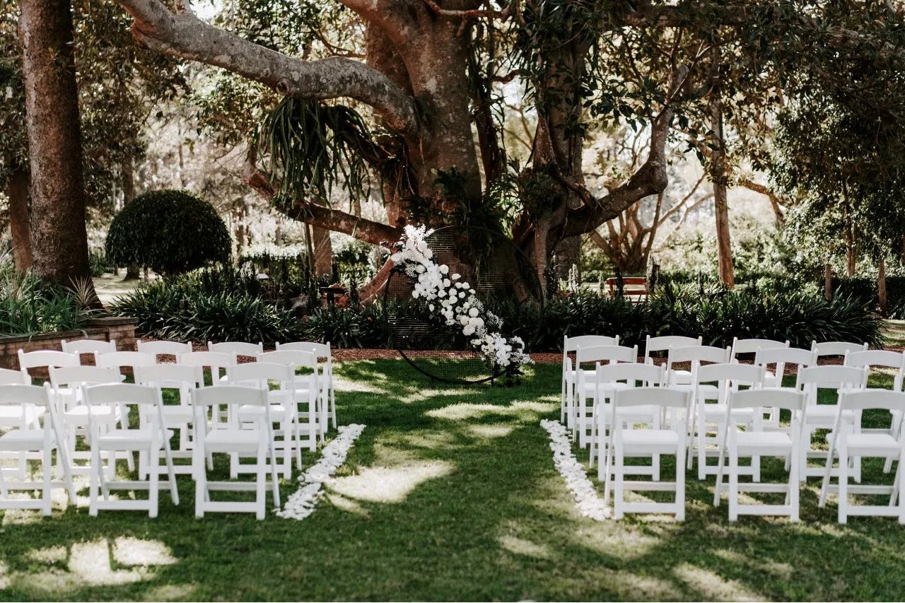 An outdoor wedding setup featuring rows of white chairs arranged on a grassy area, facing a large tree adorned with white flower decorations. The scene is surrounded by lush greenery, creating a serene and natural setting.