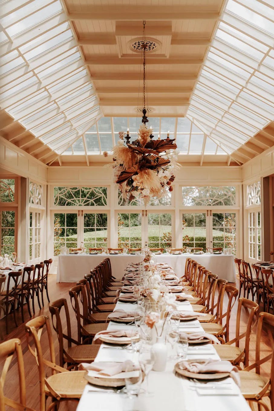 A long, elegant dining table is set for a formal meal in a bright room with large windows and a glass ceiling. The table is adorned with neatly arranged plates, glasses, and decorative centerpieces. Wooden chairs line both sides of the table, and greenery is visible outside.