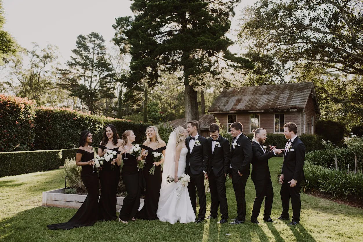 A wedding party stands outdoors on a green lawn in front of a rustic wooden shed. The bride and groom are in the center; the bride wears a white gown, and the groom a black suit. Bridesmaids in black dresses and groomsmen in black suits flank the couple, smiling and laughing.