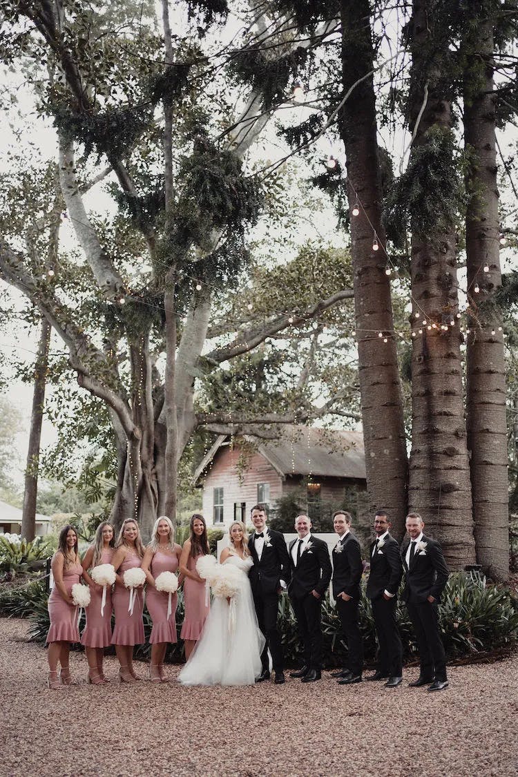 A wedding party poses outdoors among tall trees and string lights. The bride is in a white dress and veil, while bridesmaids in pink dresses hold white bouquets. The groom and groomsmen are in black suits. They all stand on a gravel path with greenery and a rustic house in the background.