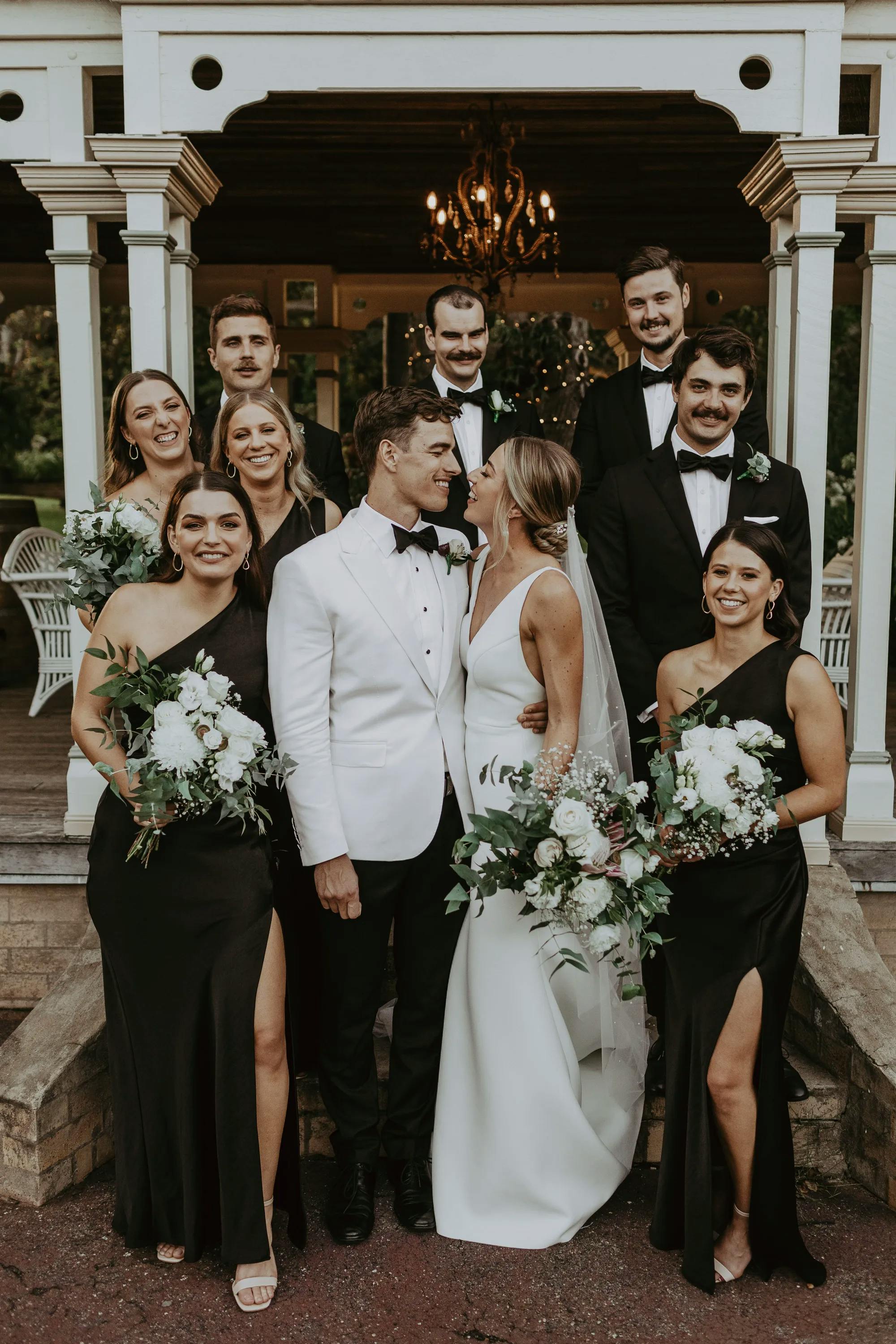 A newlywed couple stands in front of a group of smiling bridesmaids and groomsmen. The bride wears a white gown and veil, while the groom dons a white tuxedo jacket. The bridesmaids wear black dresses with bouquets, and the groomsmen are in black tuxedos. They are on the steps of a wooden gazebo.