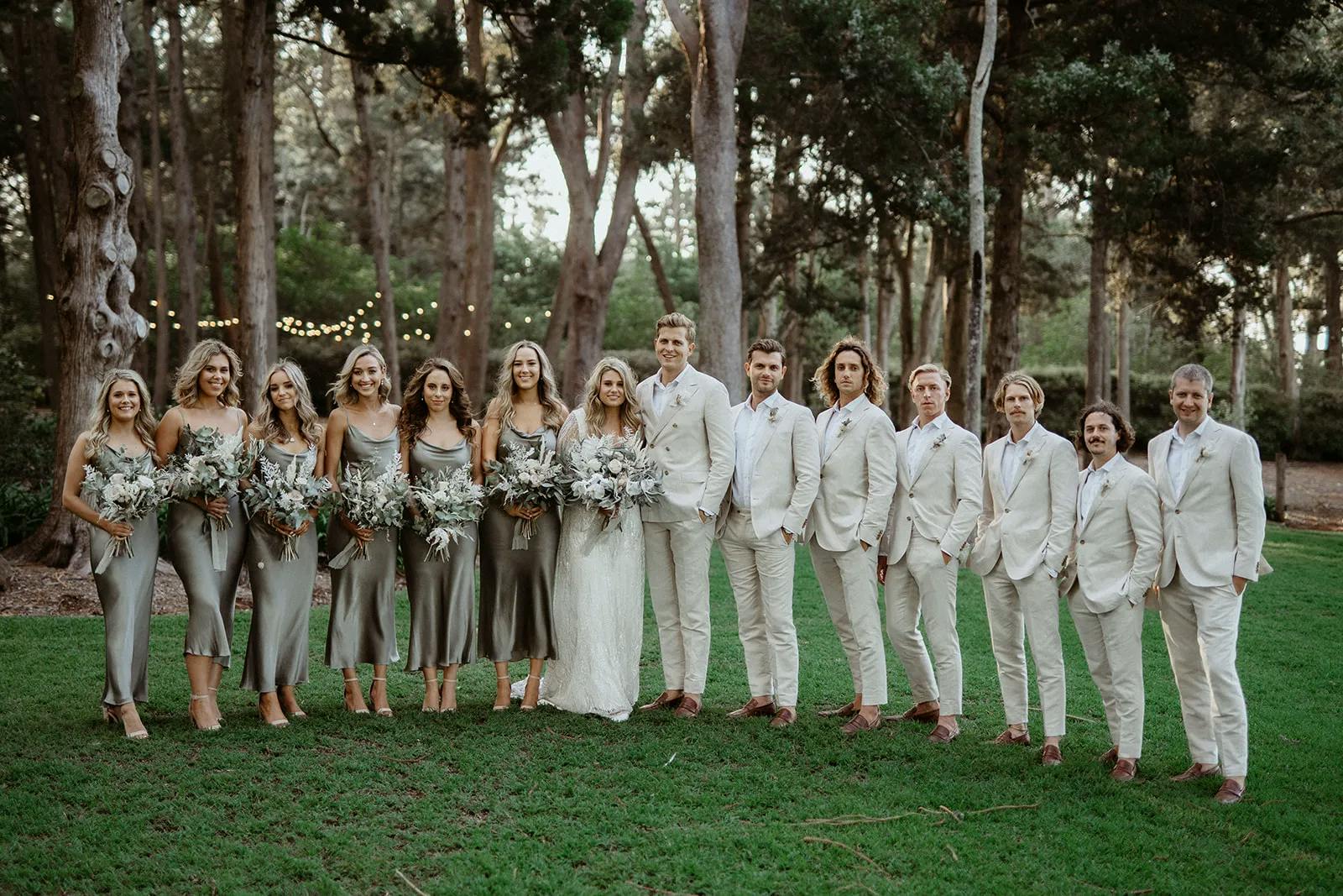 A wedding party stands outdoors on green grass with a backdrop of trees and string lights. The bridesmaids wear matching silver dresses, and the groomsmen wear light beige suits. The bride, in a white gown, and the groom, in a matching suit, stand in the center.
