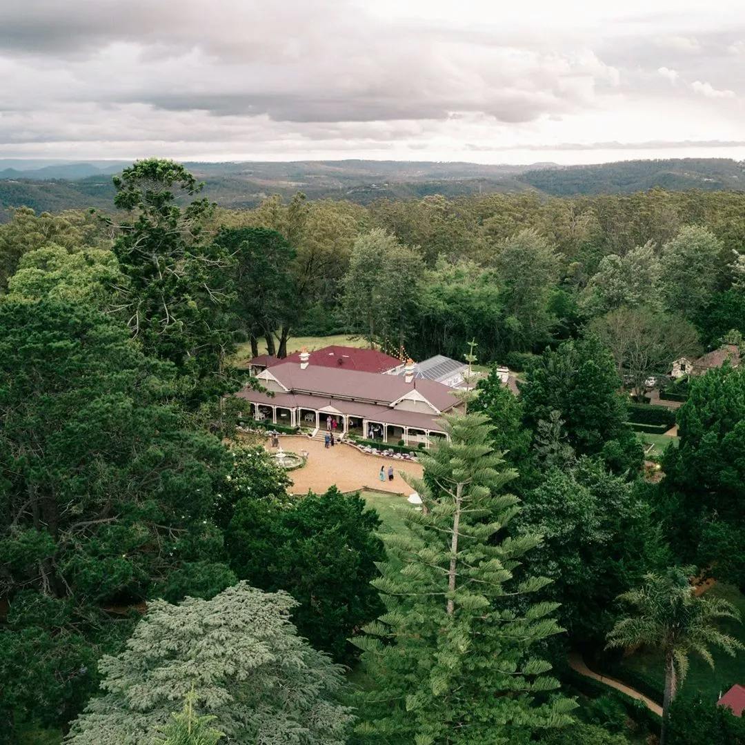 Aerial view of a large house with a red roof surrounded by dense green trees and rolling hills in the distance. The property includes a circular driveway and manicured gardens, under a cloudy sky. People can be seen near the entrance of the house.