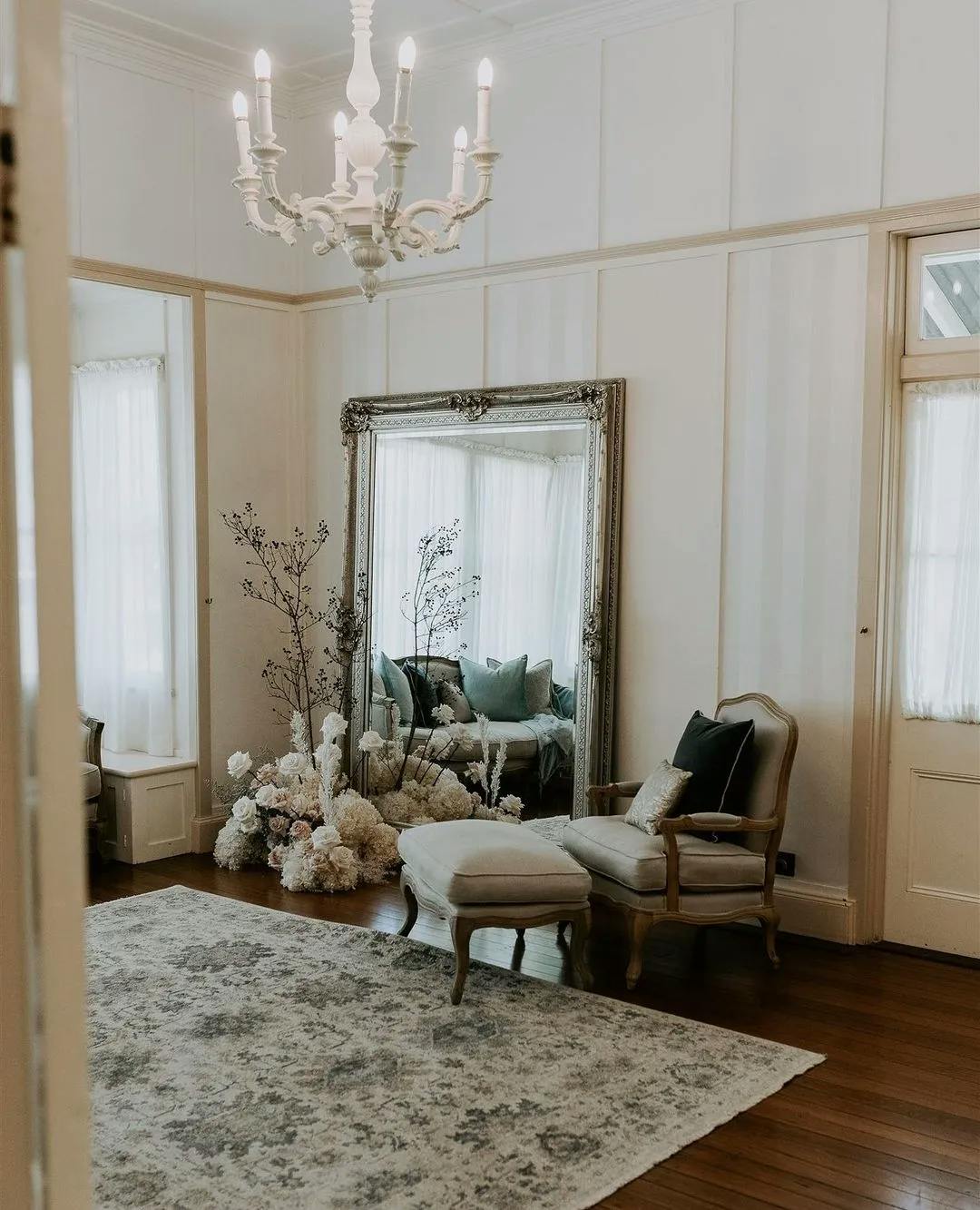A bright, elegant room featuring a large ornate mirror, flowers arranged at its base, and a white chandelier hanging from the ceiling. An upholstered armchair with an ottoman is positioned in front of the mirror on a plush rug, with large windows providing natural light.