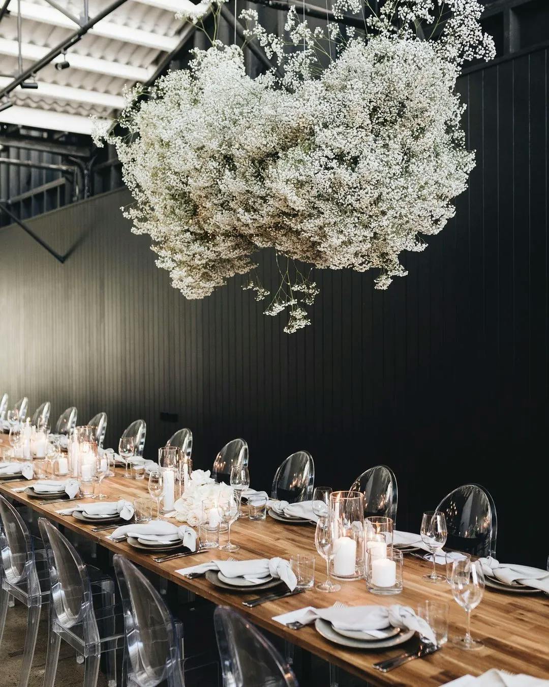 A long wooden table is set for a meal with white plates, napkins, glassware, and candles. Clear chairs surround the table. Above, a large, lush cloud-like arrangement of baby's breath flowers is suspended. The walls are dark, contrasting with the bright setting.