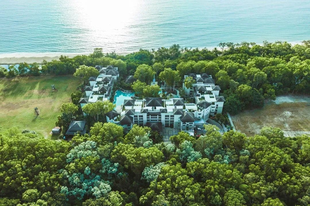 An aerial view of a beach resort surrounded by lush greenery. The resort features multiple buildings with a central swimming pool, and stretches along a sandy coastline with calm ocean waters. Dense forestation encircles the property on three sides.