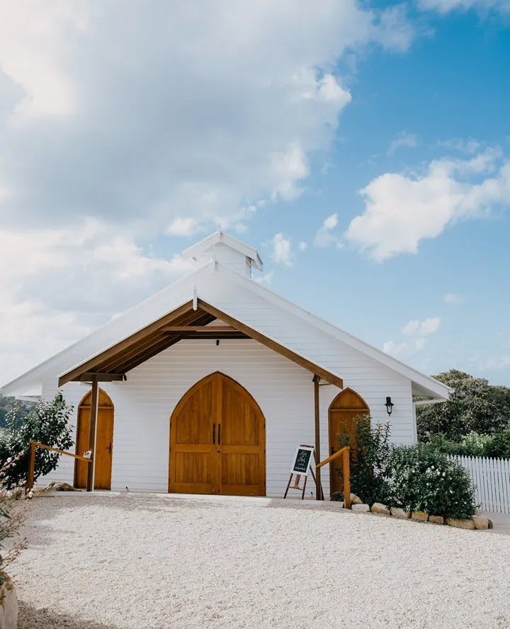 A small, white chapel with wooden doors and simple architectural features stands against a bright, partly cloudy sky. The chapel has a rustic signboard at the entrance, surrounded by shrubs and a gravel pathway leading up to it.