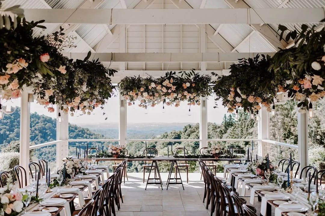 A beautifully decorated outdoor wedding reception under a white pavilion with a mountain view. The long tables are adorned with floral arrangements and greenery. Large flower arrangements hang from the ceiling. Rows of wooden chairs line both sides of the tables.