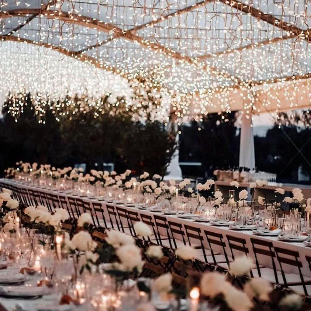 A beautifully decorated outdoor banquet table is set for a special event. White flowers and candles line the length of the table, which is illuminated by numerous hanging string lights. The ambiance is elegant and romantic, perfect for an evening celebration.