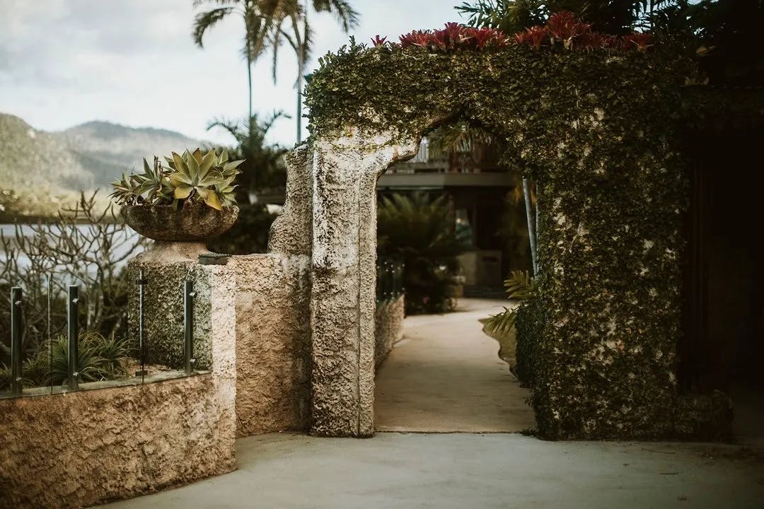 A stone archway covered in green vines and red flowers leads to a paved pathway with potted plants on a stone ledge. The background features palm trees and mountainous scenery under a partly cloudy sky.