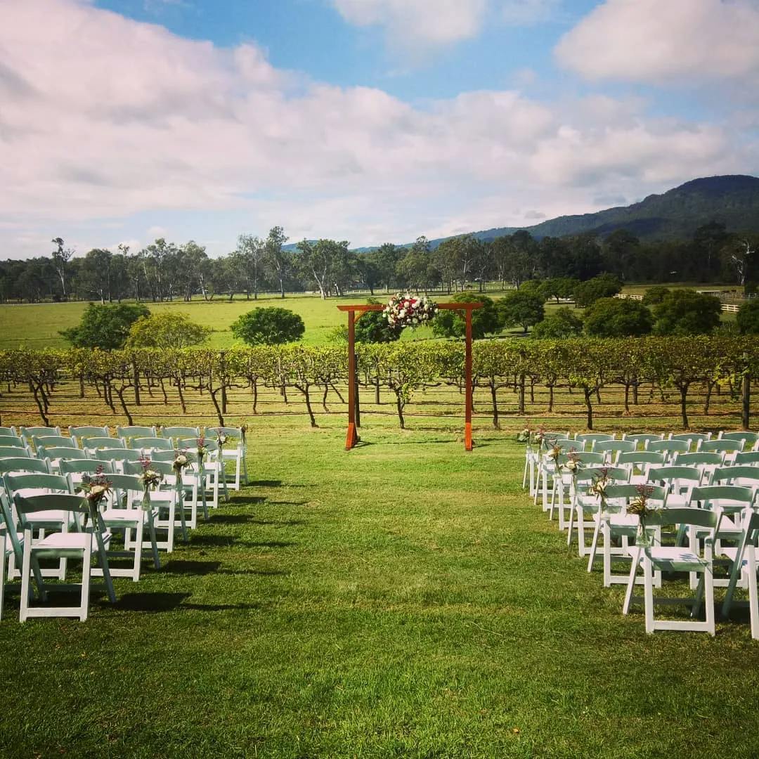 An outdoor wedding ceremony setup in a vineyard with rows of white chairs facing a wooden arch adorned with flowers. The background features lush green grapevines, trees, and rolling hills under a partly cloudy sky.