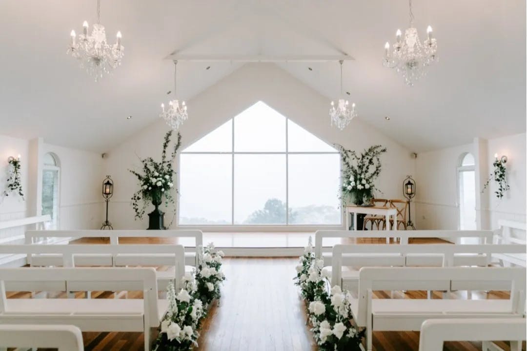 A bright and elegant wedding chapel with large windows at the front, allowing natural light to flood the room. White benches are aligned in rows, an aisle is decorated with white flowers, and chandeliers hang from the ceiling, enhancing the sophisticated ambiance.