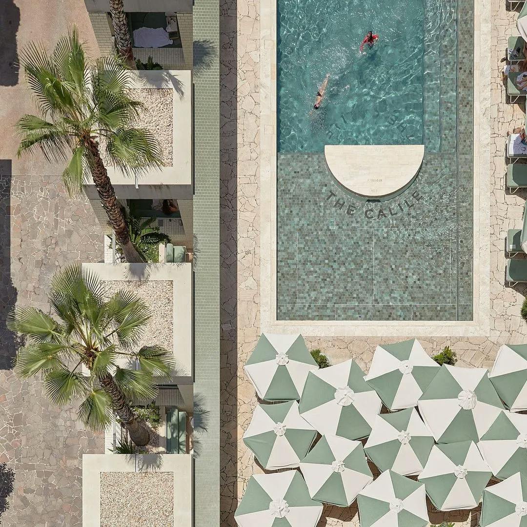 Aerial view of a swimming pool area at a resort. Two people are swimming in the pool, which has "THE CAULI" text partially visible on its floor. Surrounding the pool are sun loungers and green-and-white umbrellas. Palm trees line the left side of the image.