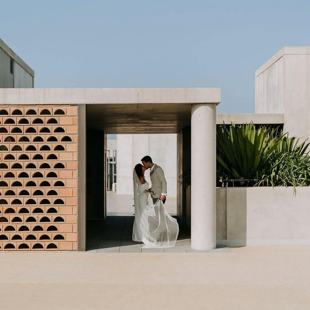A couple is standing close together in front of a modern architectural structure. The bride is in a white wedding dress, and the groom is in a light suit. They are surrounded by geometrical and green plant designs, with a clear blue sky in the background.
