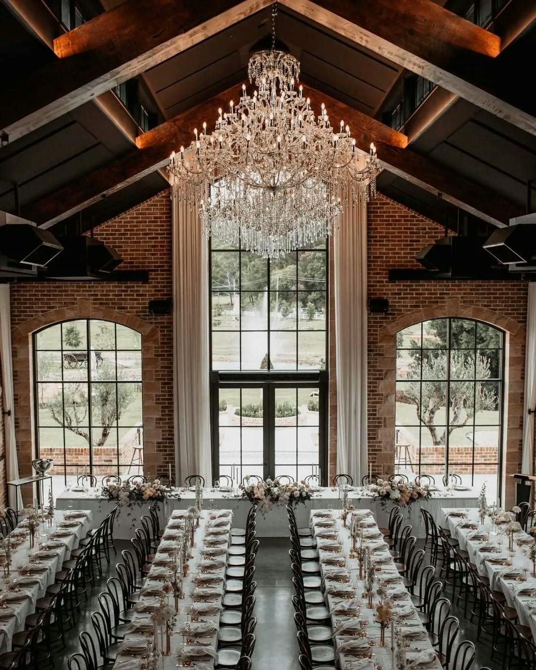 A beautifully decorated reception hall with long, white-draped tables arranged in parallel rows, adorned with floral centerpieces. Overhead, a large, ornate chandelier hangs from the high wooden-beamed ceiling. Tall windows let in natural light.