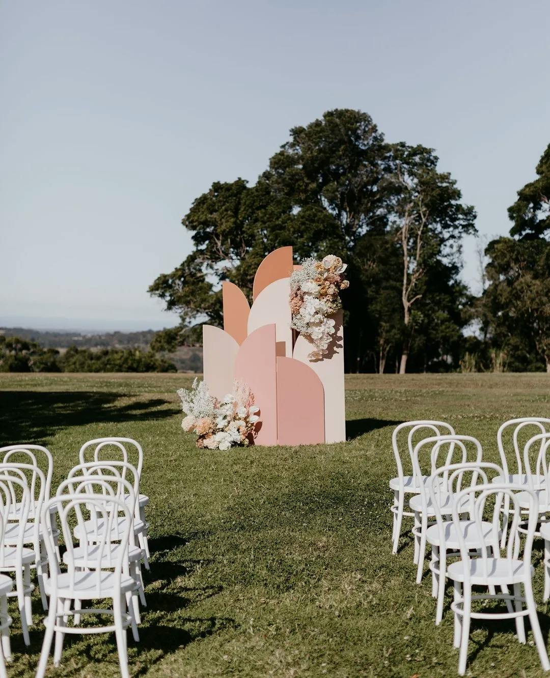 An outdoor wedding setup with white chairs arranged in rows on grass facing a pink and orange geometric backdrop adorned with floral arrangements. Tall trees and a distant landscape are visible in the background.