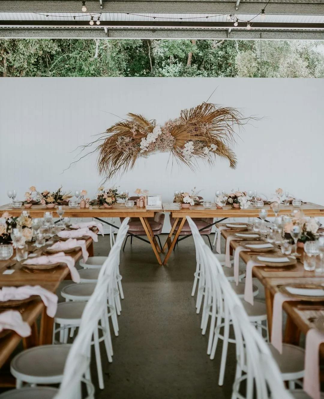 A beautifully set dinner table in an outdoor setting with white chairs and wooden tables. The tables are decorated with pink napkins, flowers, and glassware. A white wall with a rustic dried floral arrangement is visible in the background.