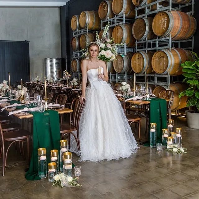 A bride in a white wedding dress stands holding a glass of champagne in a rustic venue with wooden barrels stacked on one side. The tables are elegantly set with green table runners, floral arrangements, and candles in glass vases on the floor.