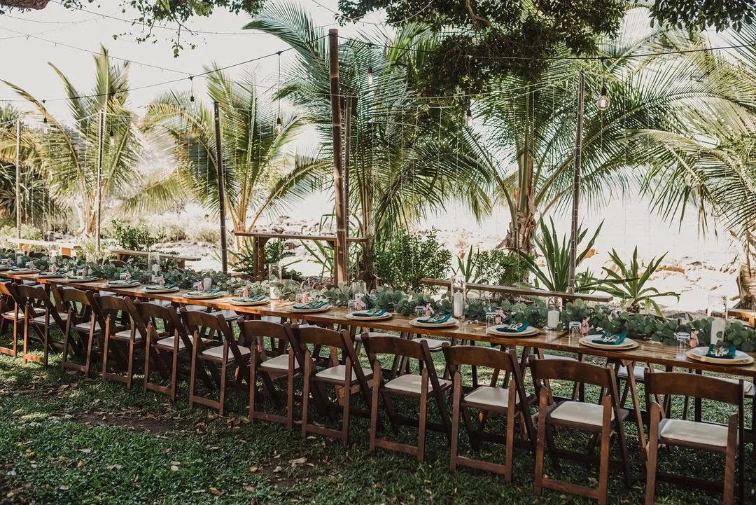 A long rectangular outdoor dining table is set up with green plates and decorative foliage, surrounded by wooden chairs. The setup is nestled in a tropical garden with lush palm trees and hanging string lights, creating a serene and elegant ambiance.