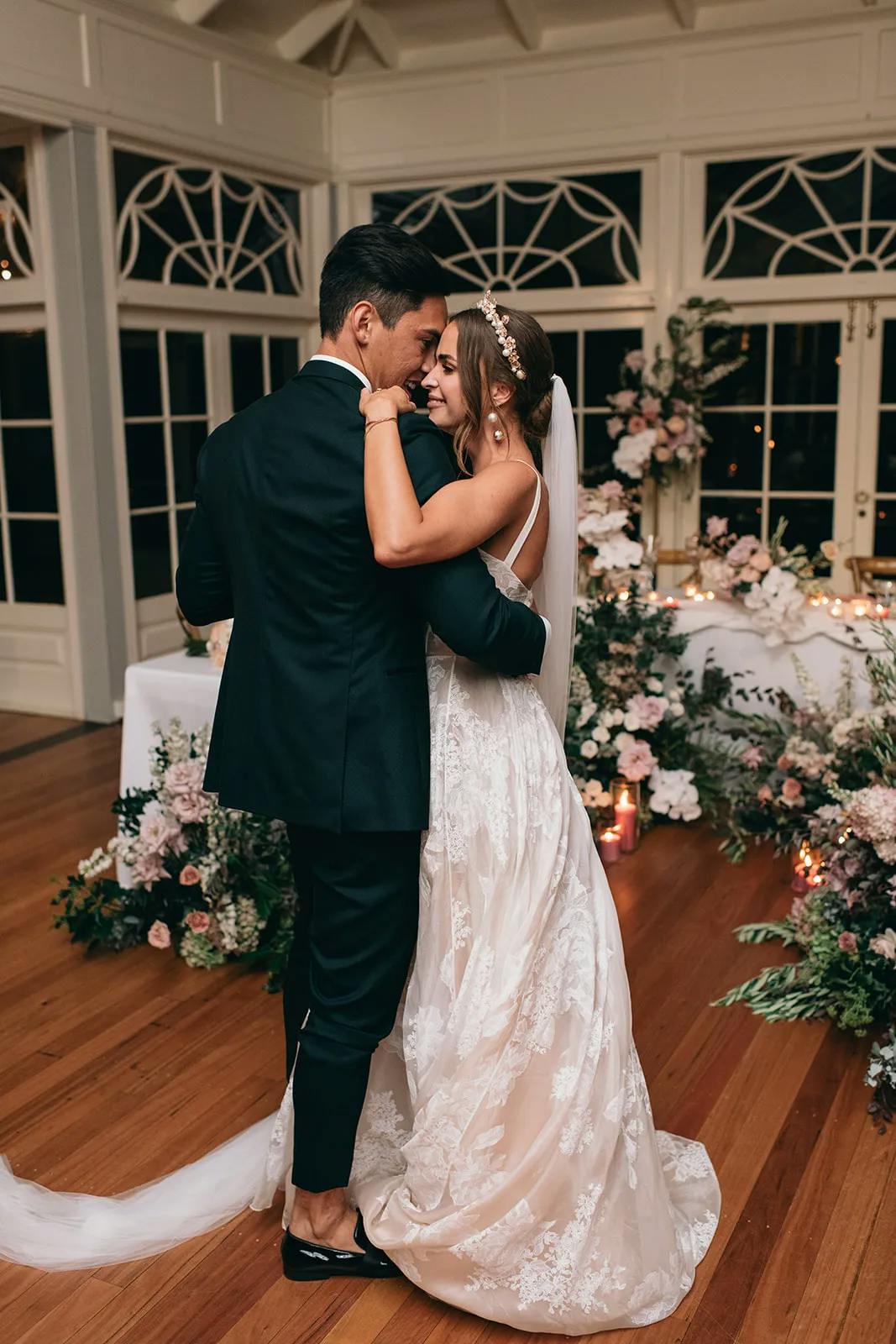 A bride and groom share a dance at their wedding reception. The bride, in a white lace gown and veil, smiles as she embraces the groom, who is wearing a black suit. The venue is elegantly decorated with floral arrangements and soft lighting, creating a romantic atmosphere.
