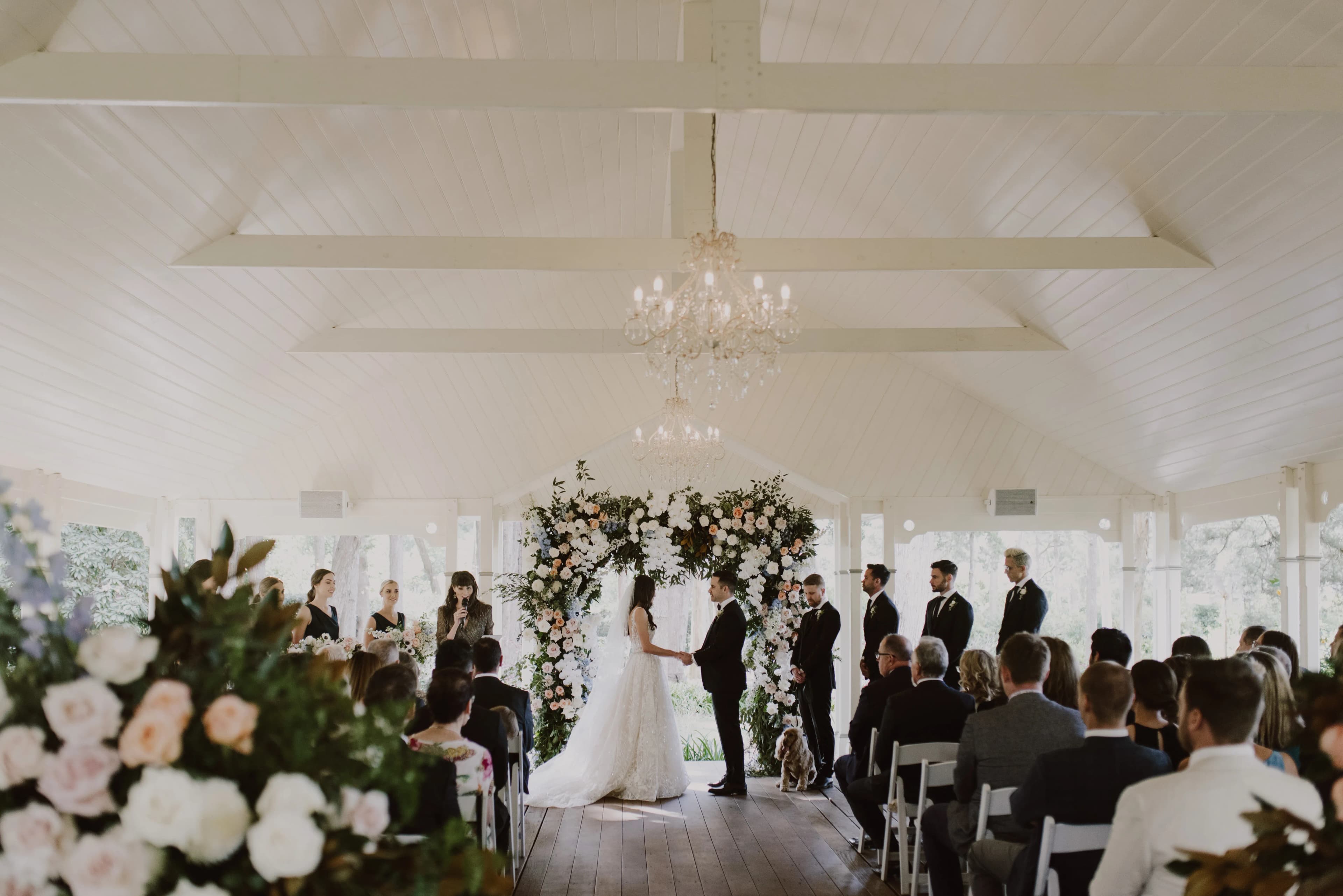 A couple stands facing each other under a floral arbor, exchanging vows in a bright, white pavilion. Guests sit in rows, attentively watching the ceremony. The space is adorned with elegant chandeliers, and large floral arrangements frame the aisle.