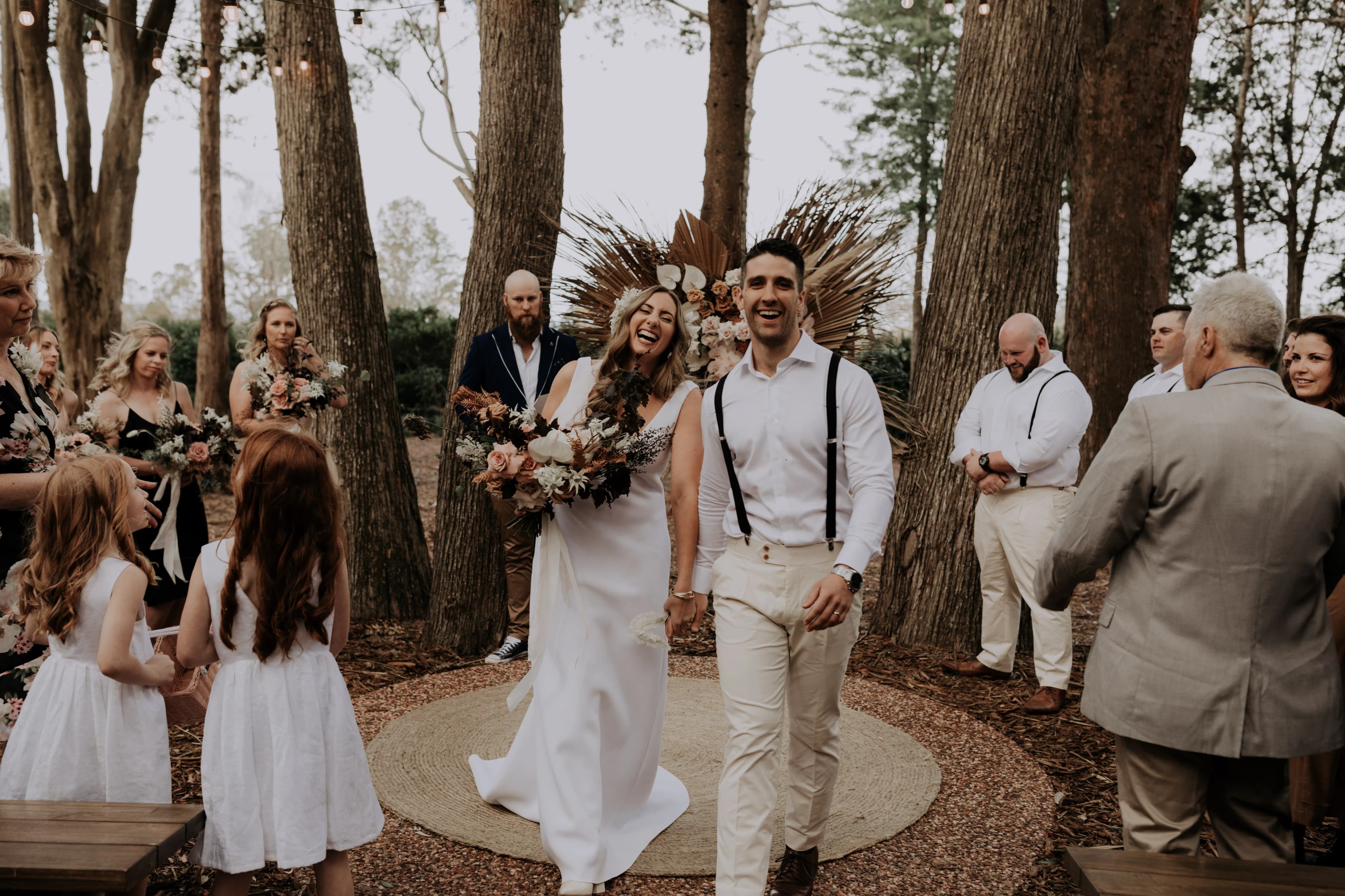 A bride and groom, both smiling and holding hands, walk down an aisle outdoors surrounded by tall trees. The bride, in a white gown and floral headpiece, laughs joyfully. The groom in white attire with suspenders looks elated. Guests in casual attire look on happily.