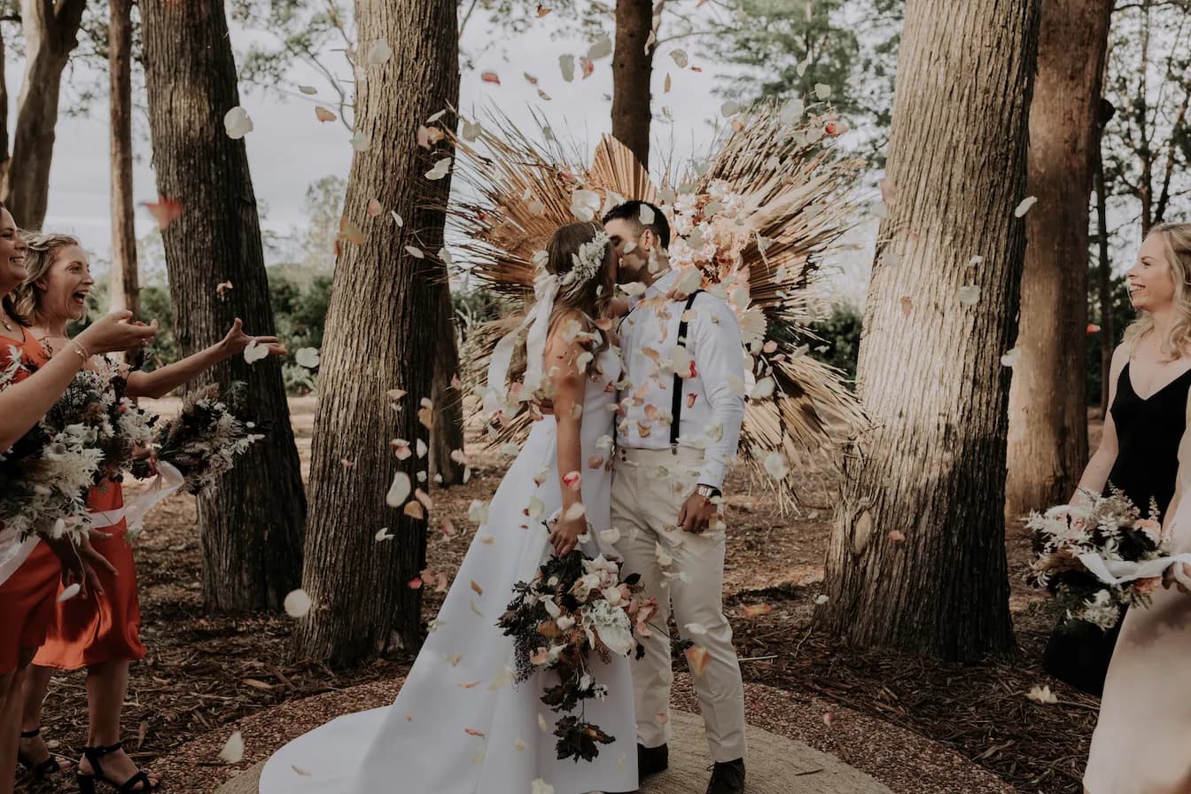 A bride and groom share a kiss outdoors among tall trees, surrounded by friends throwing flower petals. The bride wears a white dress and holds a bouquet, while the groom wears light-colored attire. A decorative arrangement of dried plants forms the backdrop.