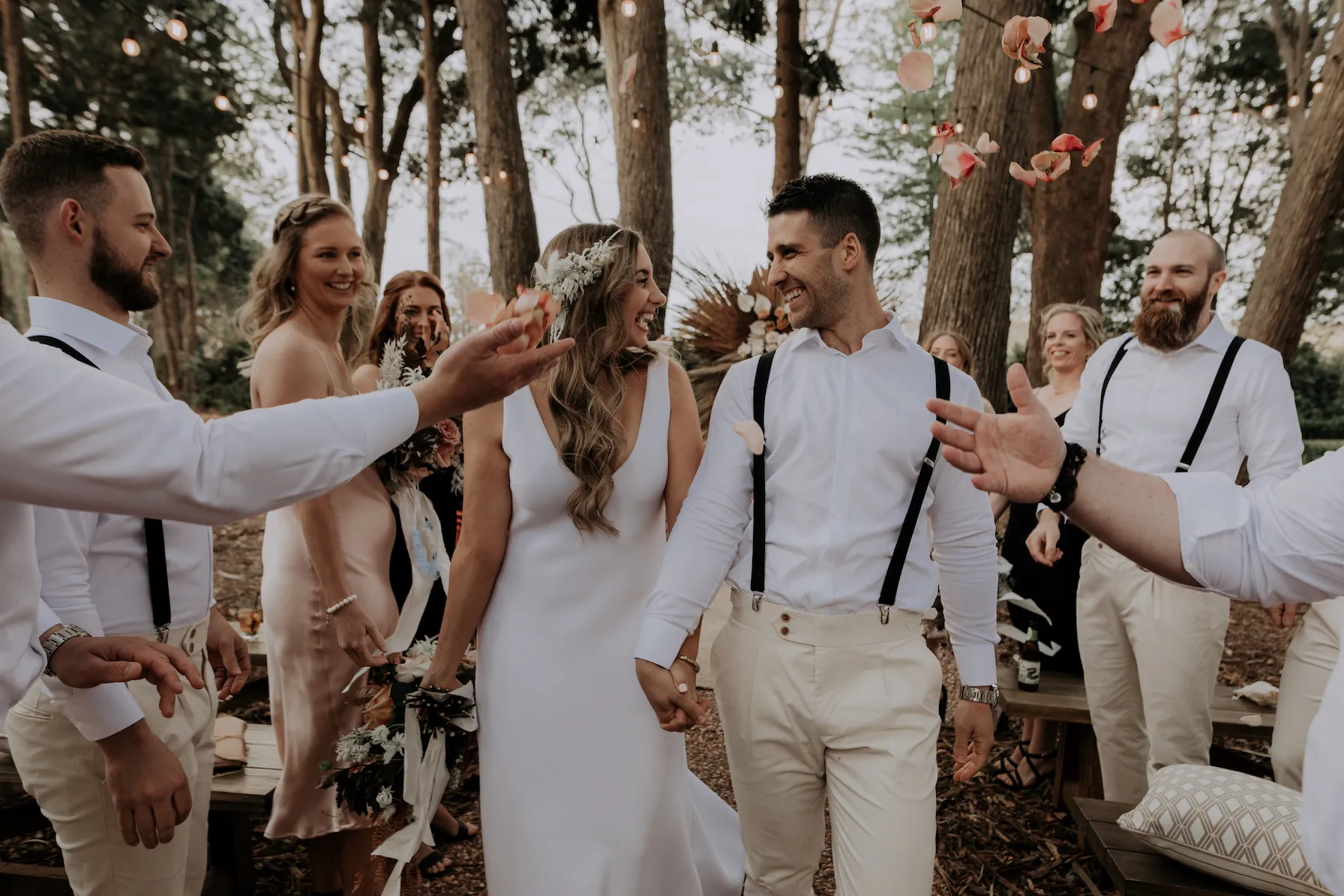 A newlywed couple walks hand-in-hand down an outdoor aisle surrounded by joyous friends and family. The group is dressed in light-colored attire, and the setting is a wooded area with string lights and scattered floral decorations. The atmosphere is celebratory and warm.