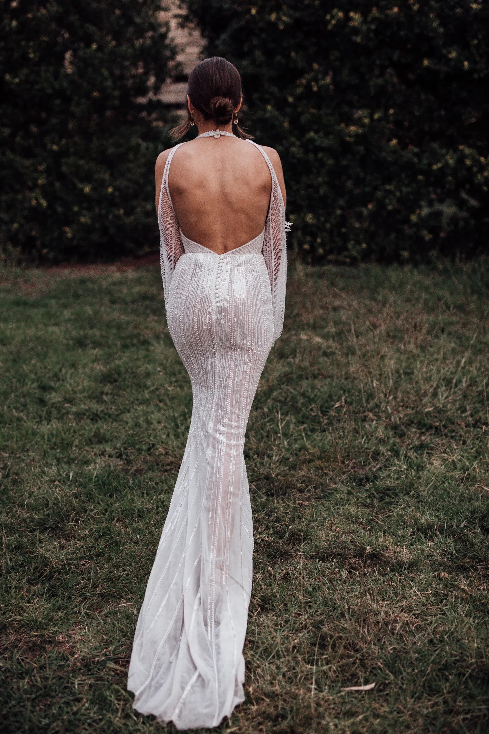 A woman stands on grass, wearing a backless, shimmering white gown with thin straps. Her hair is styled in an updo, and she faces away from the camera with lush greenery in the background.