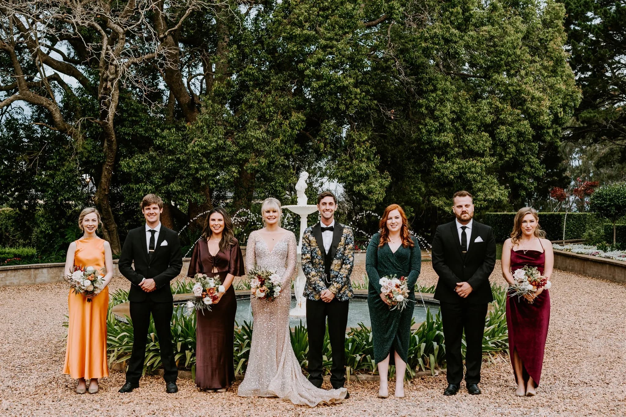 A wedding party stands in front of a lush, green backdrop with a modest fountain. The bride, in a sparkly white dress, is in the center next to the groom in a patterned tuxedo. Flanked by bridesmaids in various colored dresses and groomsmen in black suits.
