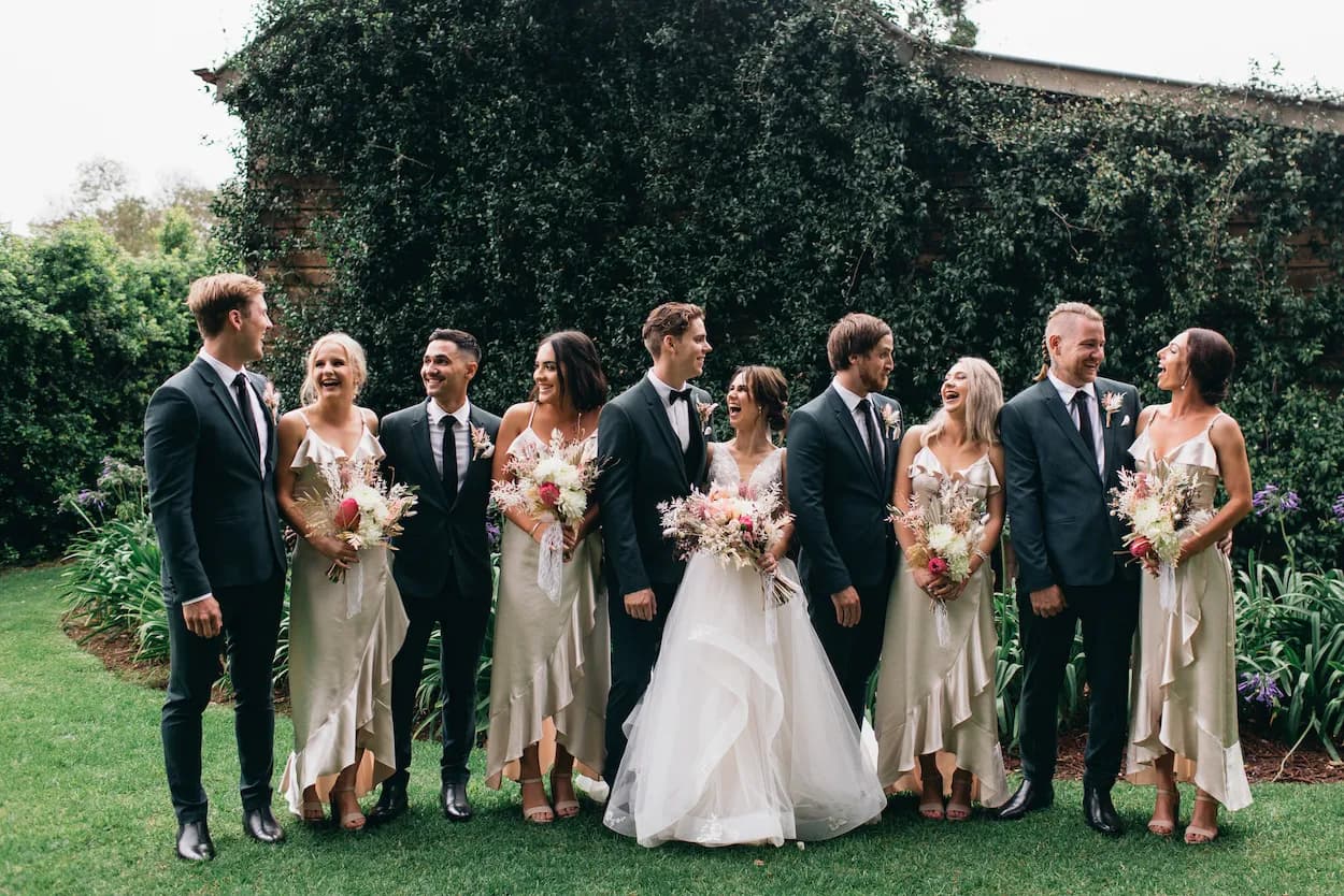 A wedding party poses outdoors on a grassy area in front of a leafy backdrop. The bride and groom stand in the center, surrounded by six bridesmaids and groomsmen, all wearing coordinated formal attire. Everyone is smiling and interacting joyfully.